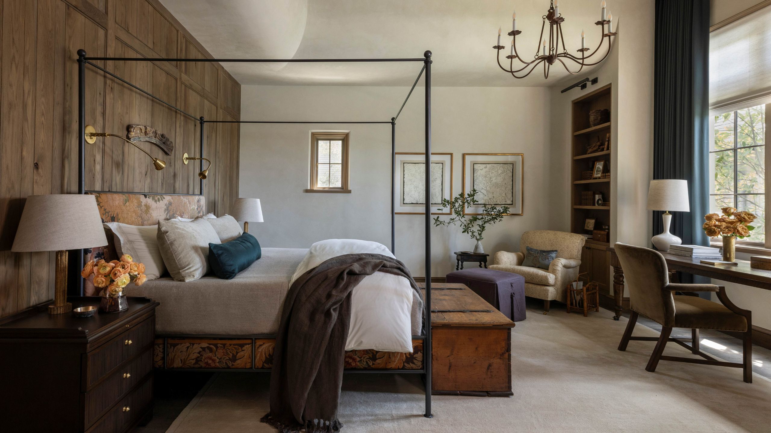 a grand primary bedroom with 1 wood clad paneled wall, a four poster tapestry wrapped bed, layered furnishings, a desk, and sitting area