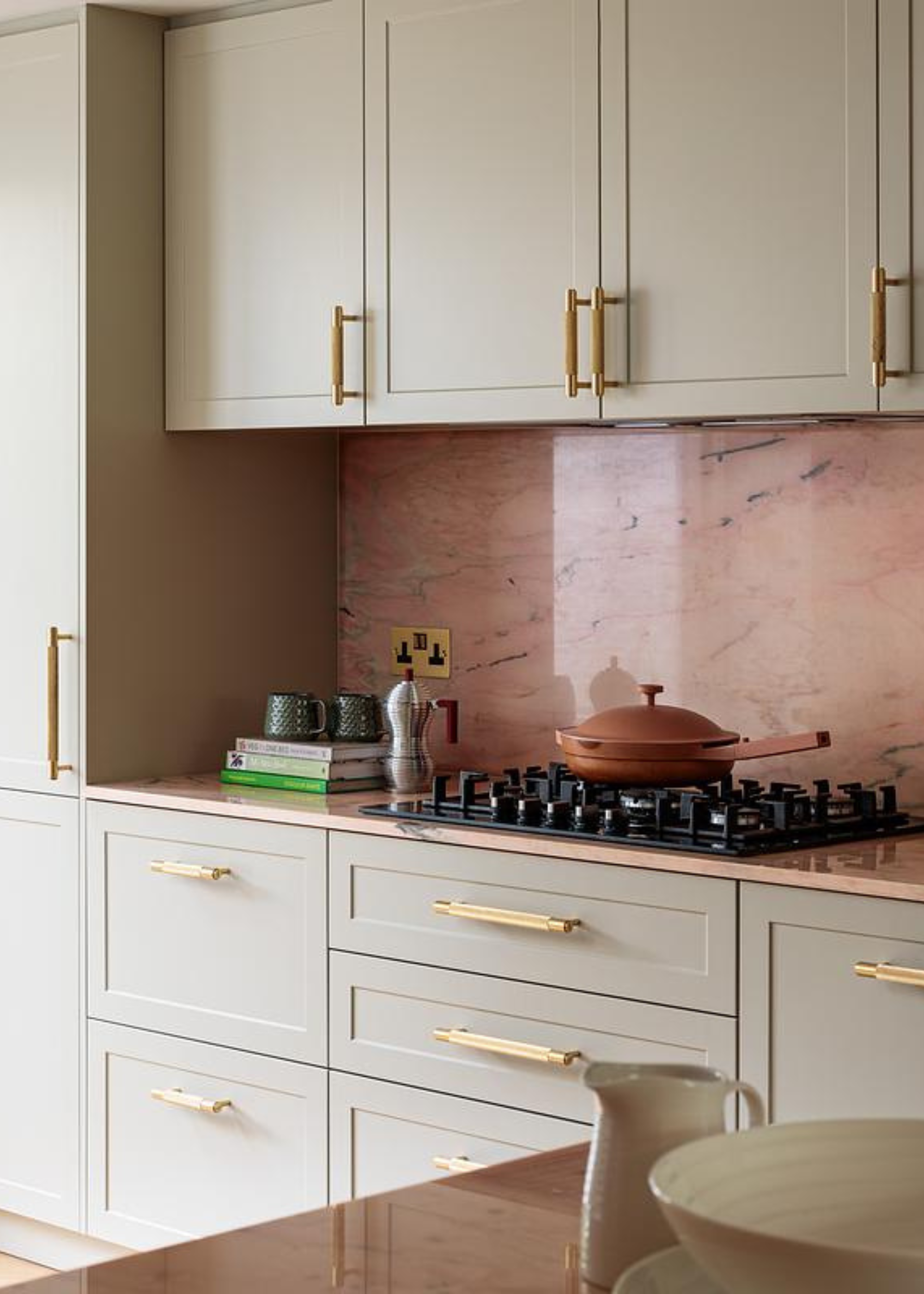 A small kitchen with a pink stone backsplash, beige cabinetry with gold hardware and a gas stove. A terracotta-colored pan with a lid, an Alessi coffee pot and a stack of books are also on display.