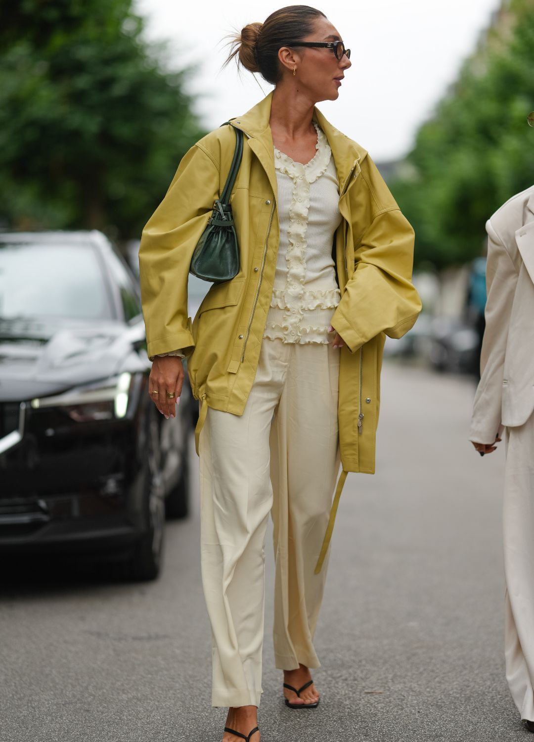 A guest wears a light olive green oversized jacket with a hood and snap button closures. Underneath, a cream-colored textured top with ruffled detailing is visible. The outfit includes cream-colored wide-leg trousers. Accessories include dark sunglasses and a small dark green leather shoulder bag. Footwear consists of black strappy sandals. Hair is styled in a neat bun, outside OperaSport, during Copenhagen Fashion Week, on August 04, 2025 in Copenhagen, Denmark