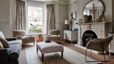 A neutral living room with a large intricate fireplace, circular mirror, white rug, mismatched sofas and couch, white chest of drawers, and bay windows with cream curtains.