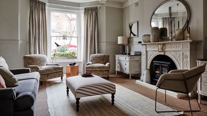 A neutral living room with a large intricate fireplace, circular mirror, white rug, mismatched sofas and couch, white chest of drawers, and bay windows with cream curtains.