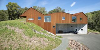 Alternative exterior view of Triple Barn house during the day. The house features three Corten steel roofs, a driveway and it is surrounded by greenery with a hill and steps beside it. There is also a car parked on the property