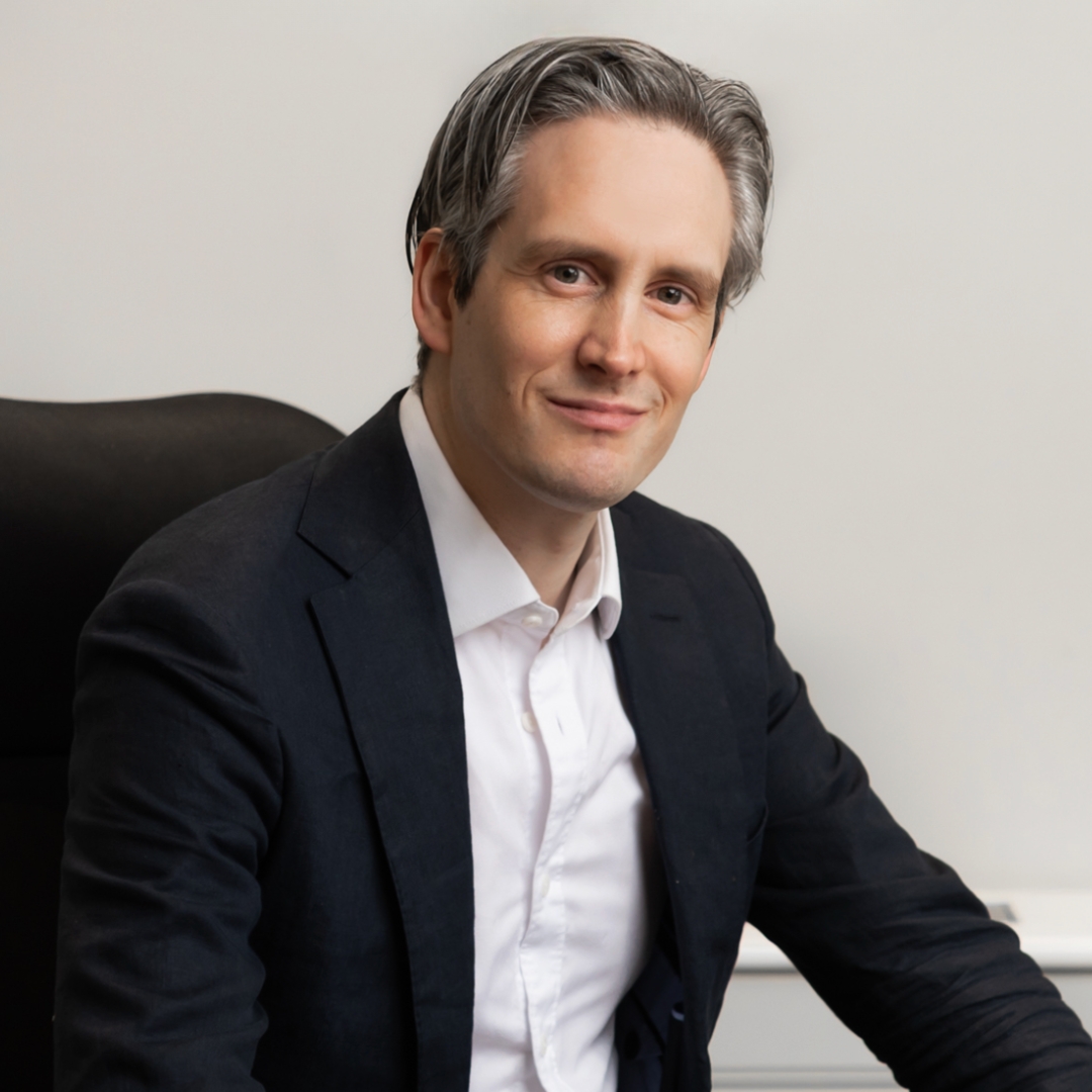 Image of Dr Magnus Lynch smiling with grey short hair and wearing a black suit blazer and white shirt