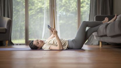 A woman dressed in workout clothes lies on a yoga mat in her living room. Her feet rest on a sofa and she gazes at a mobile phone held in her hand.