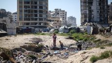 A mother and her children crossing water with tents and dilapidated buildings in the background