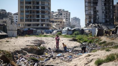 A mother and her children crossing water with tents and dilapidated buildings in the background