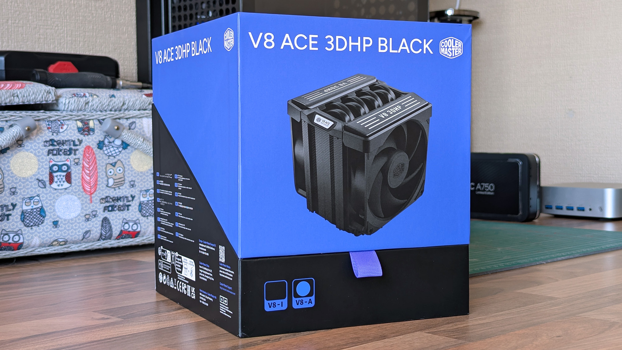 The Cooler Master V8 ACE 3DHP Black CPU cooler box sits on a wooden desk, showing the product image, branding, and specifications. Other PC components are visible in the background.