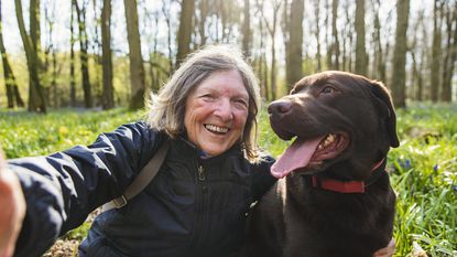 An older woman takes a selfie with her chocolate Lab dog.