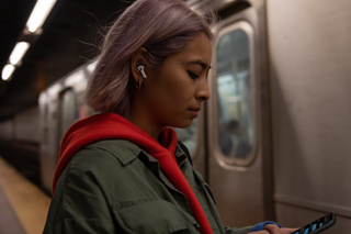 A person standing in a subway station wearing a pair of Apple AirPods Pro and looking at their phone.