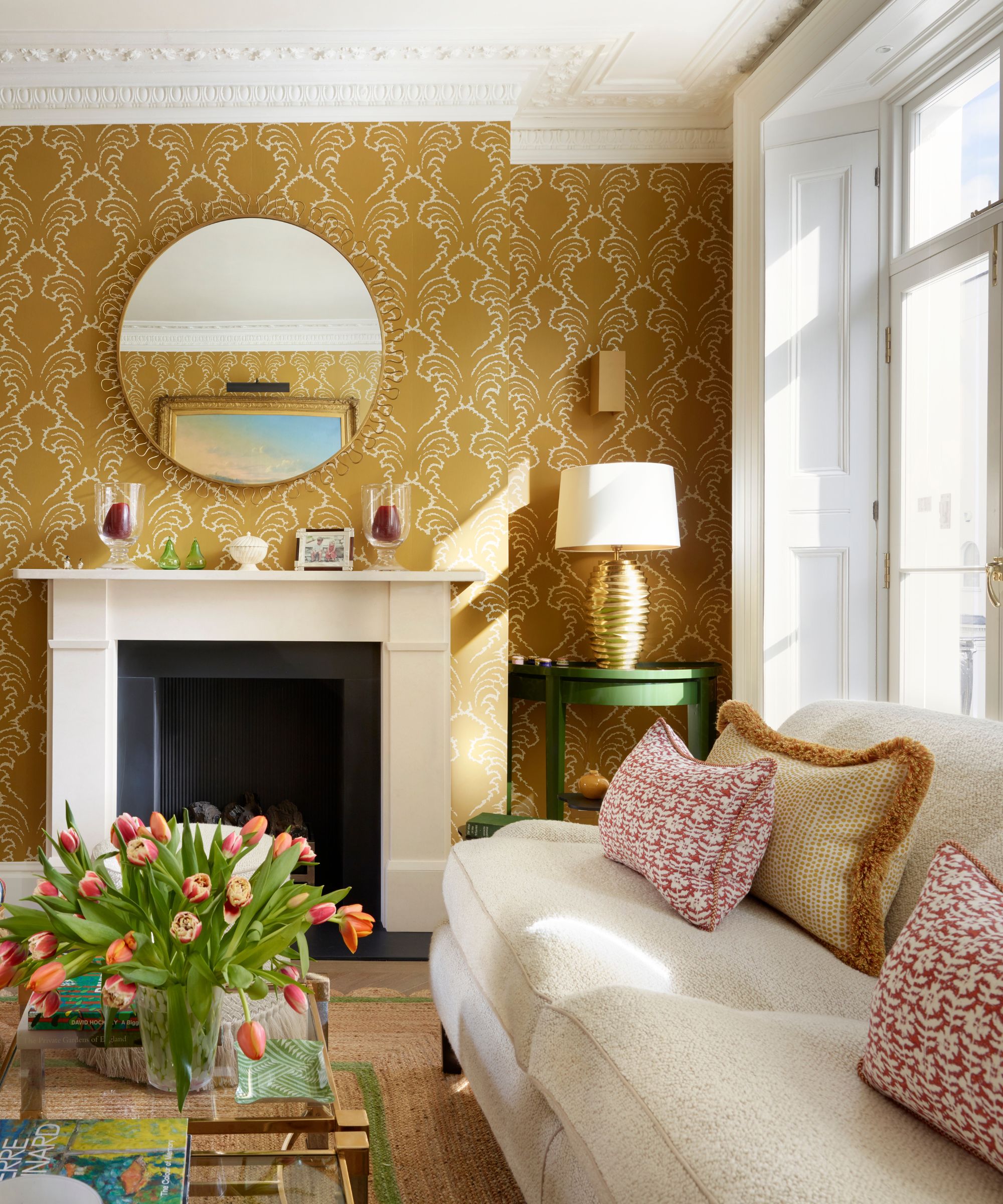 A traditional living room with ochre and white patterned wallpaper on the walls, a fireplace with a round mirror above, and a cream sofa with ochre and red cushions.