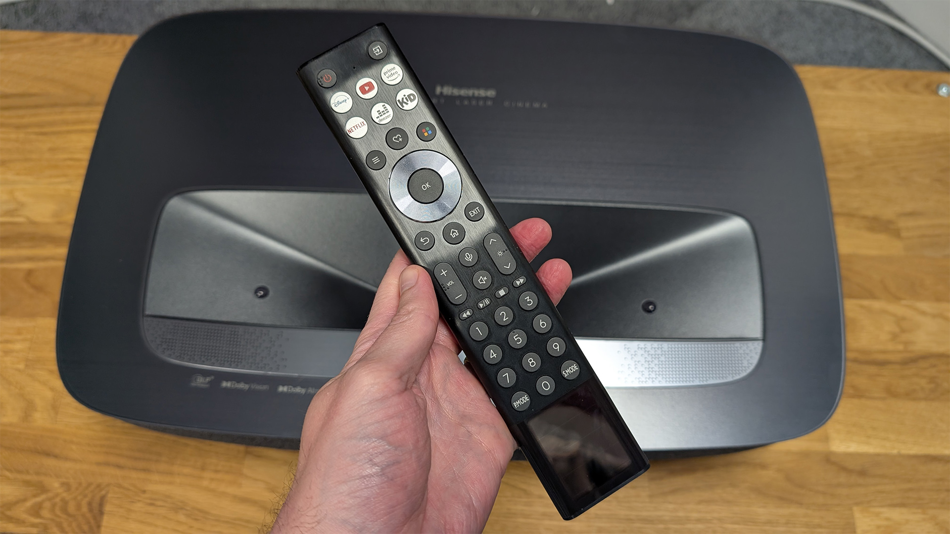 Hisense PL2 UST projector on wooden AV rack with remote control held in hand above i