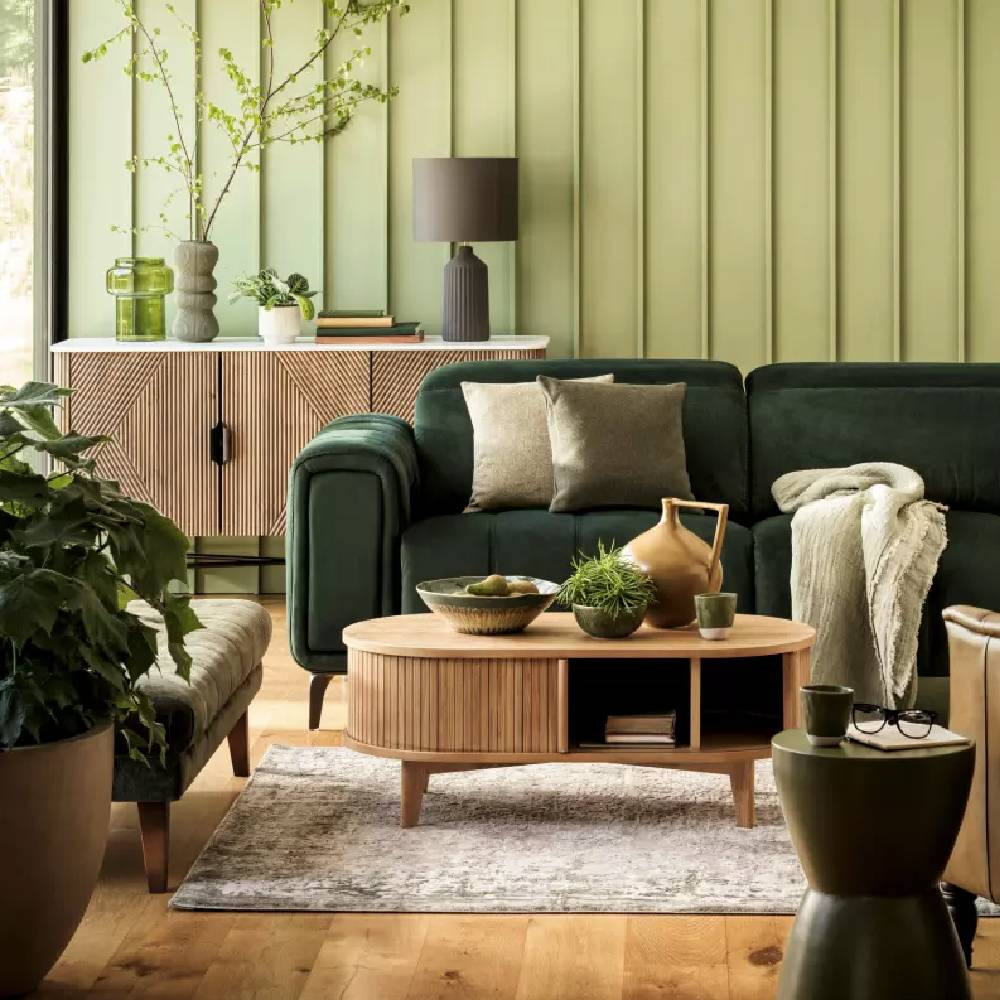 A modern, green lounge with dark, velvet sofas, a round, rattan and wooden coffee table, and various accessories, all from Furniture Village.