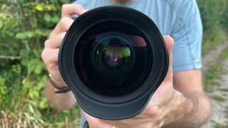 The round reflective glass of a camera lens is seen head-on, with a black lens shade around it. It is being held by two hands attached to hairy arms. The lens obscures the torso of the person with the arms.