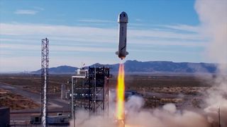 A screenshot from a YouTube video showing Blue Origin's New Shepard rocket blasting away from its launch pad against a blue sky