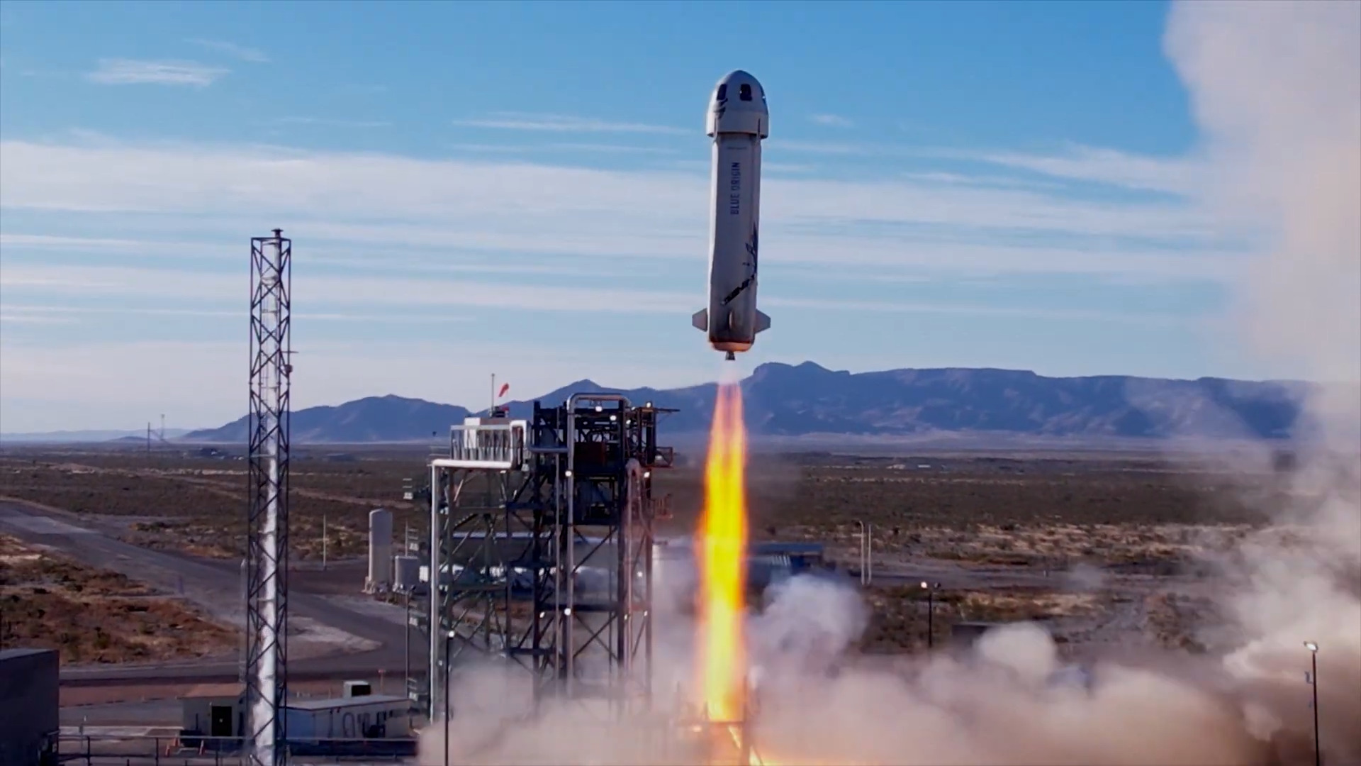 A screenshot from a YouTube video showing Blue Origin&#039;s New Shepard rocket blasting away from its launch pad against a blue sky