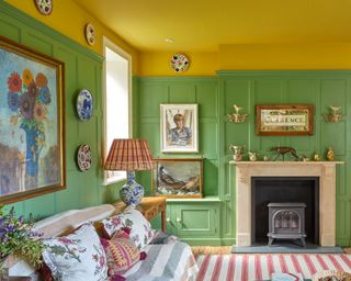 Colorful living room with yellow painted ceiling, green painted paneled walls, eclectic mix of framed artwork and pictures, decorative plates on the wall, traditional stone fireplace, red and white striped rug, striped throw and patterned cushions on sofa, blue and white ceramic table lamp with patterned shade