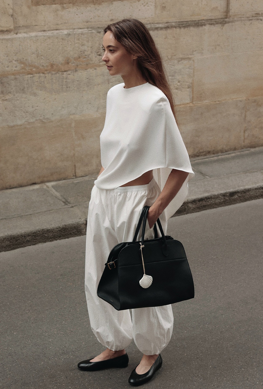 A woman in Paris wearing a white cape shirt styled with white balloon pants, black ballet flats, and a black bowler bag.