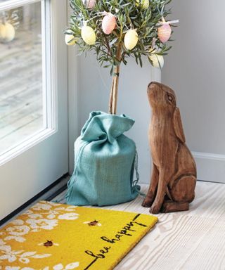 A yellow doormat that says 'bee happy' in black writing with a small Easter egg adorned tree in a blue bag next to it and a dark wooden bunny rabbit ornament next to that
