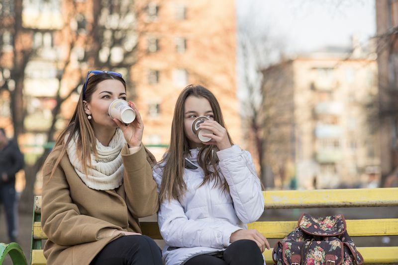 Two women sit on a bench drinking coffee