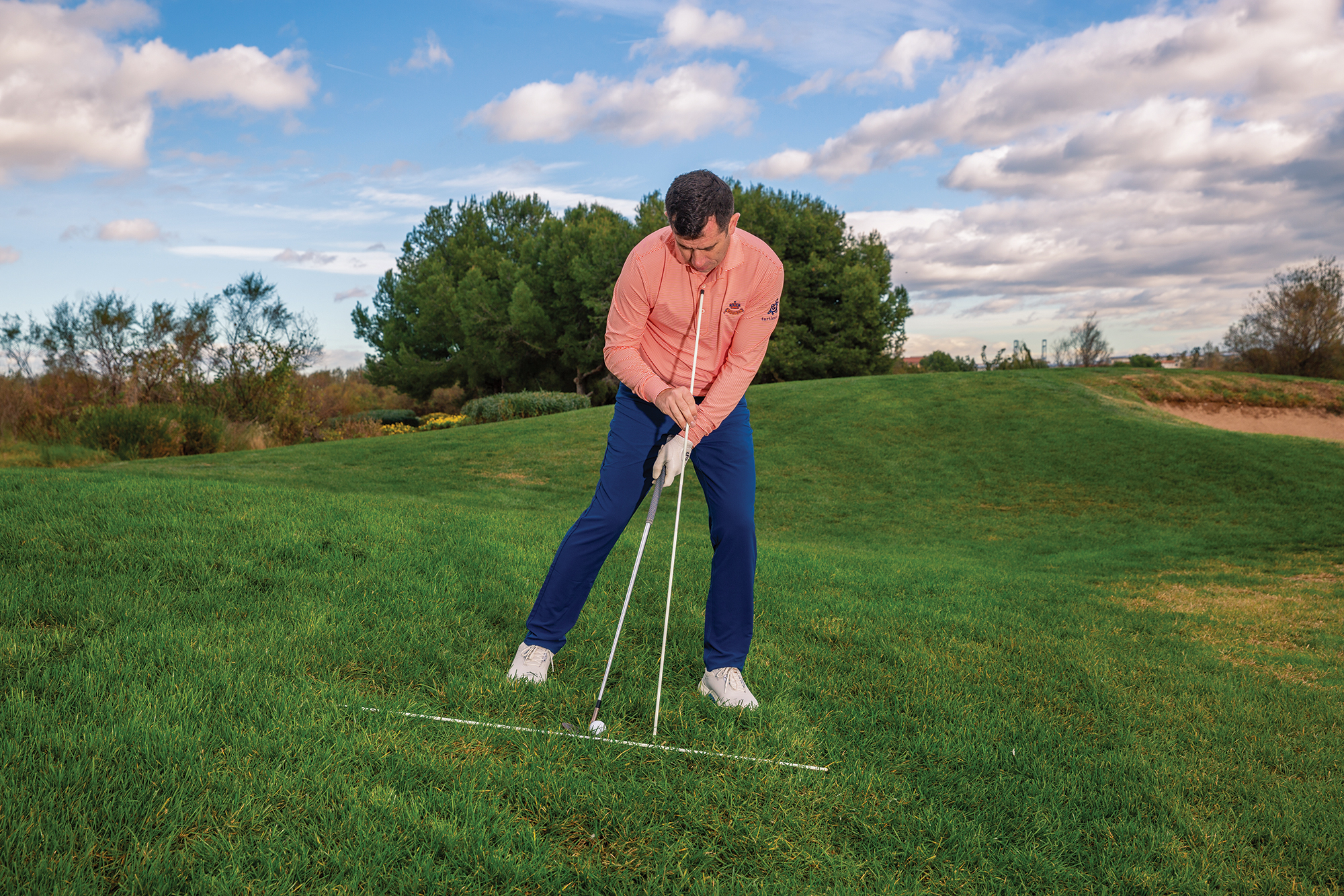 Dan Grieve using alignment sticks to demonstrate a tip on how to hit a downhill chip shot from the rough, while also holding a wedge behind the golf ball