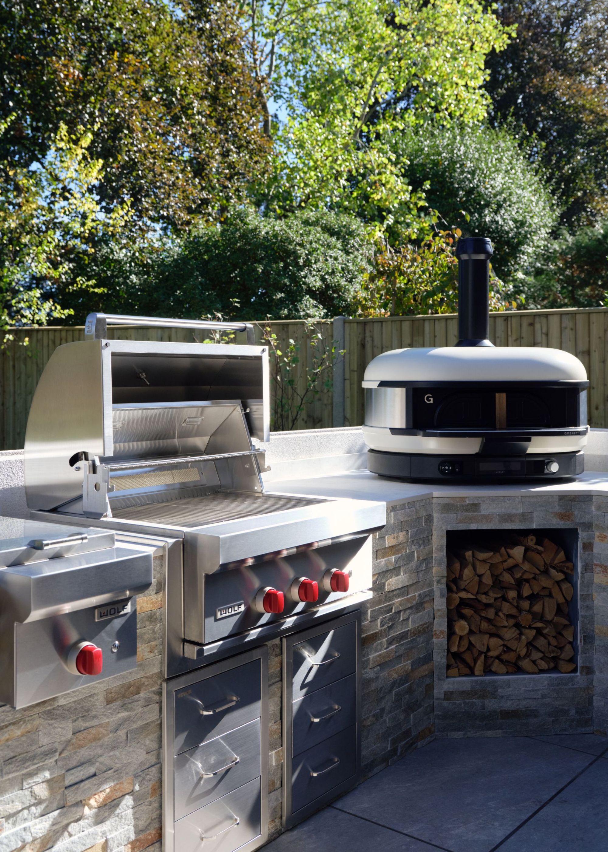 An outdoor kitchen with a gozney dome pizza oven in bone with wood underneath and a subzero &amp;amp; wolf oven with the lid opened and drawers underneath