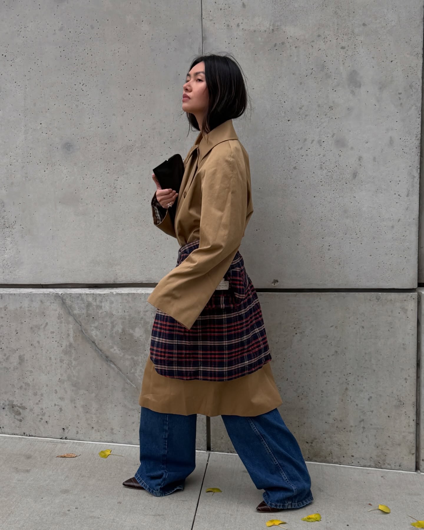 Woman wearing a plaid shirt tied around her waist over a long trench coat and jeans.