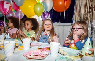 Children sitting at a table wearing party hats, surrounded by balloons, with party food on the table