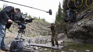 A behind-the-scenes image showing Amber Midthunder's Naru preparing to fire an arrow, with a camera crew ready to film the scene