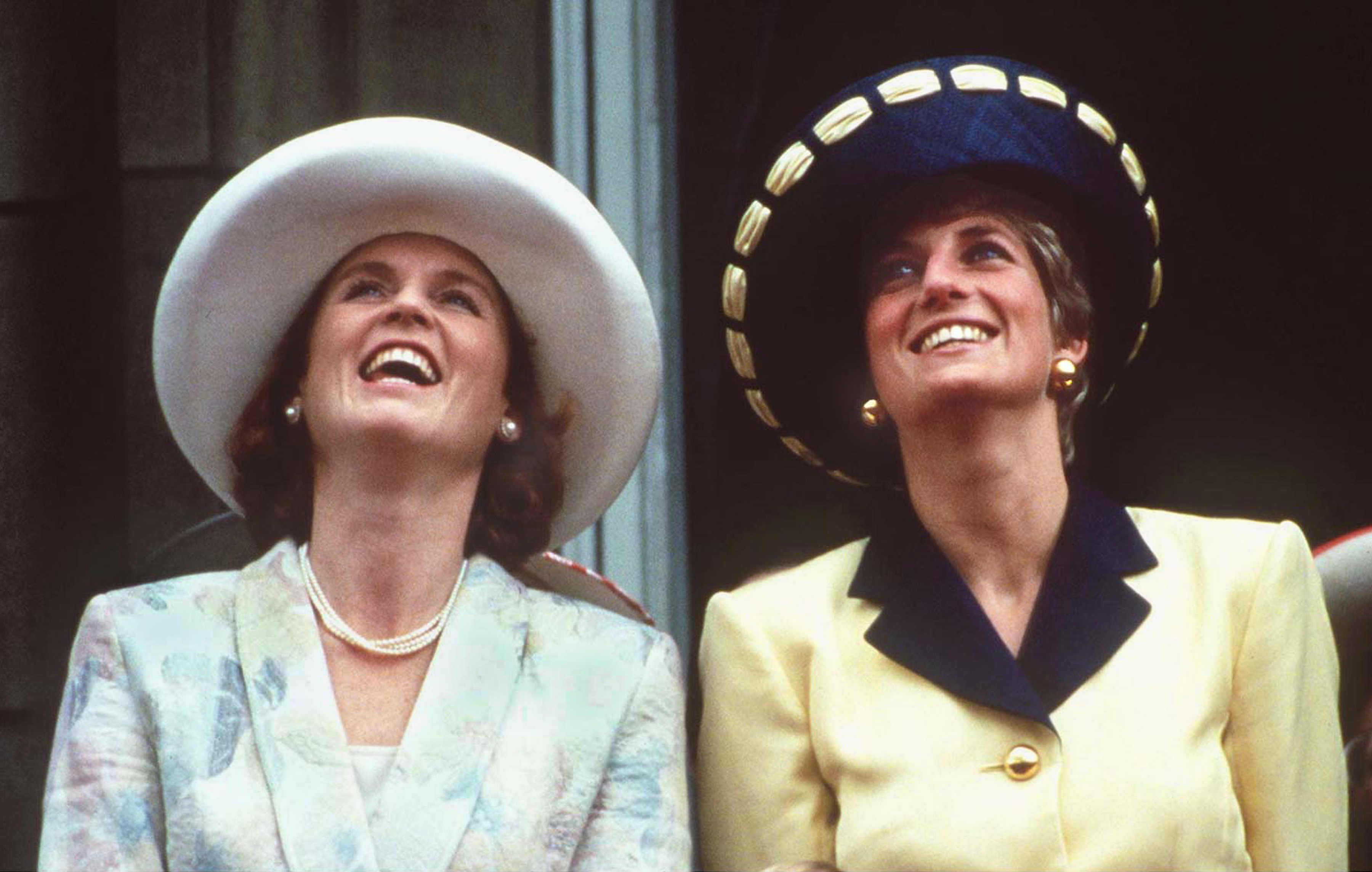 Sarah Ferguson and Princess Diana wearing hats and blazers looking up and smiling