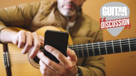 A guitarist with a nylon-string guitar in his lap holds his cellphone out in front of him