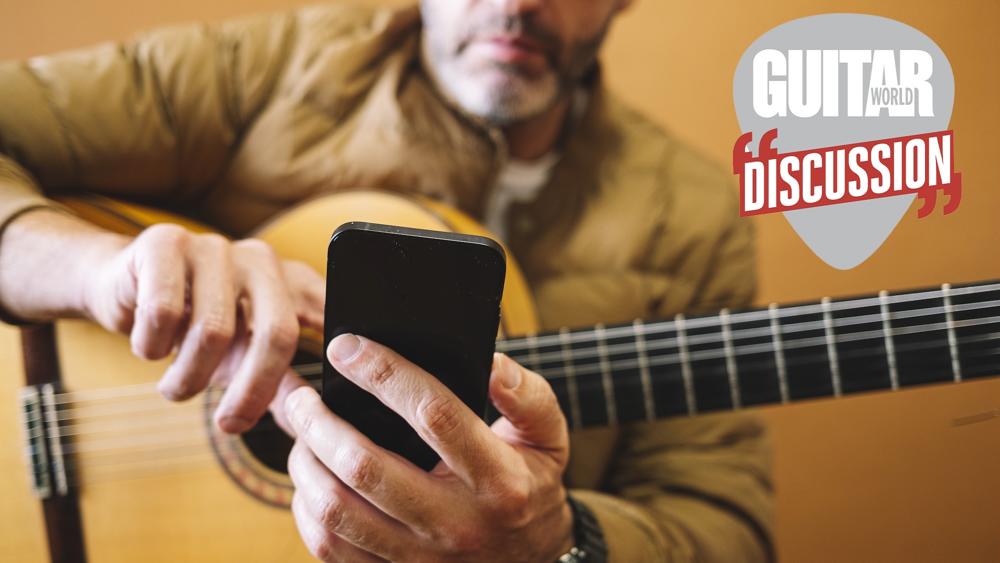A guitarist with a nylon-string guitar in his lap holds his cellphone out in front of him