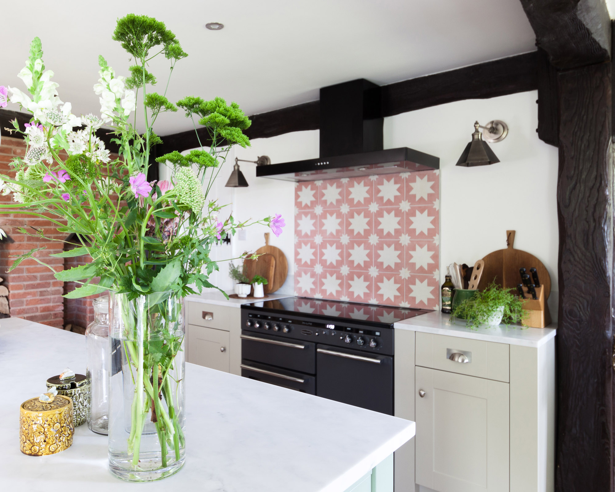 A white kitchen with a pink patterned tile backsplash, black oven and large vase of flowers in the foreground.