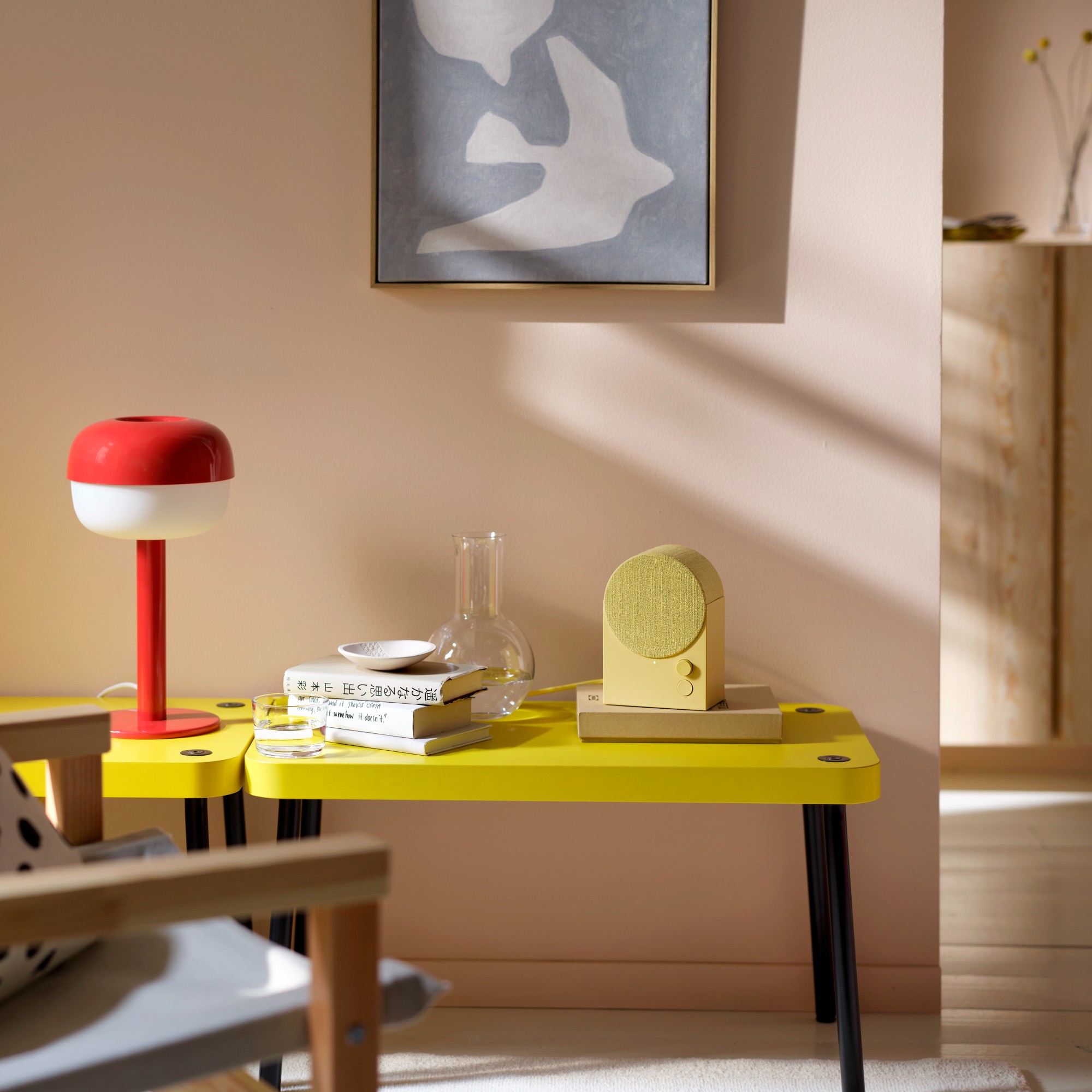The IKEA NATTBAD bluetooth speaker in yellow on a yellow side table against a pink wall and next to a red IKEA table lamp 