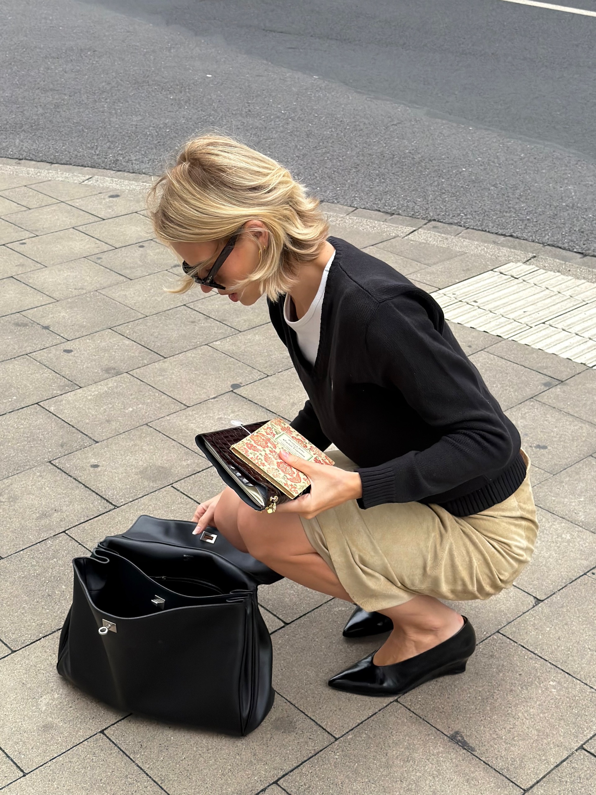 An image of an influencer wearing vintage-inspired micro wedge heels, an autumn shoe trend, with a beige skirt and black V-neck knit putting items into a large black tote