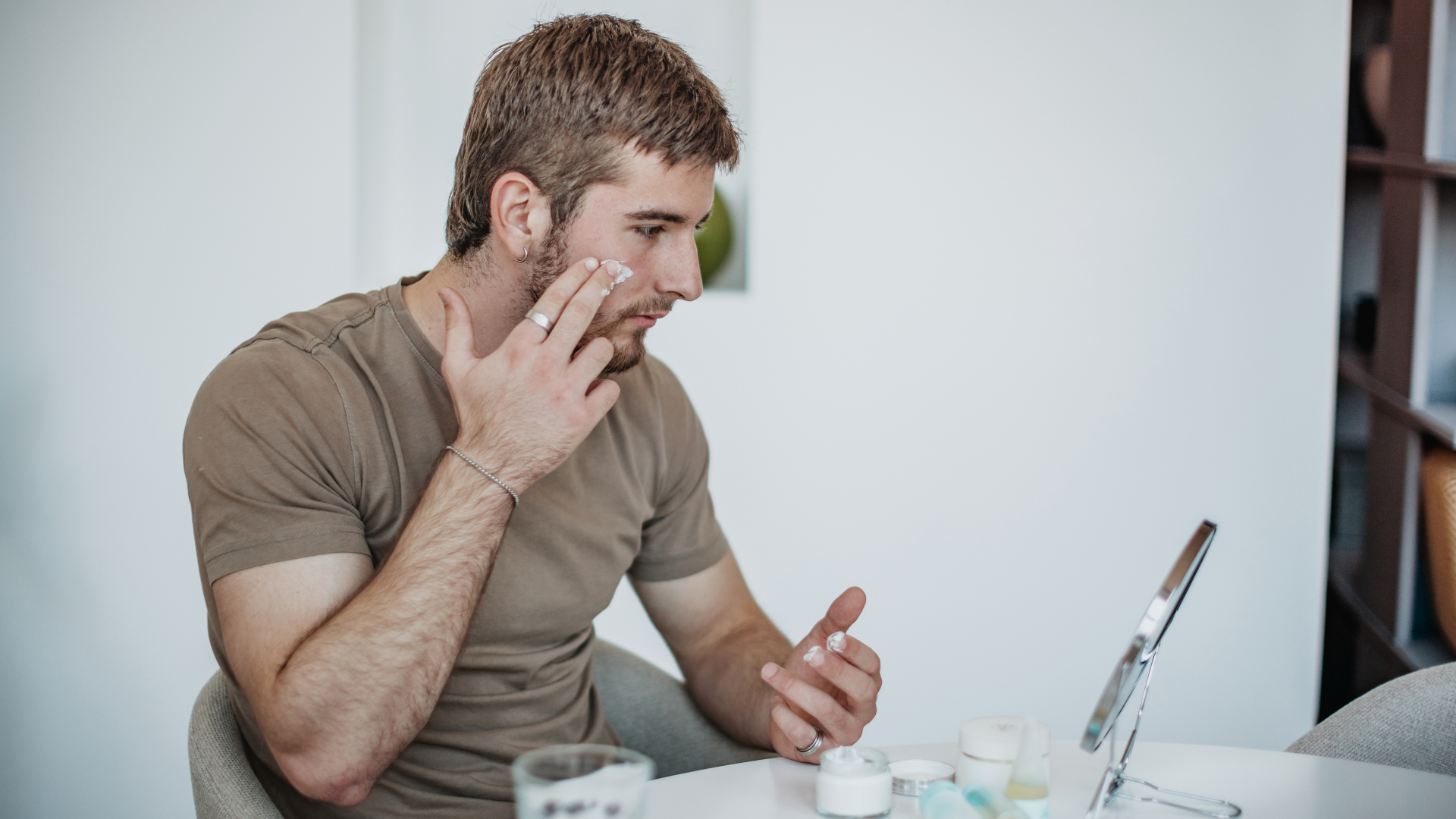 A man is sitting at a table in his home. He is applying skin care cream to his face using his fingers. Various skin care products are laid out in front of him.