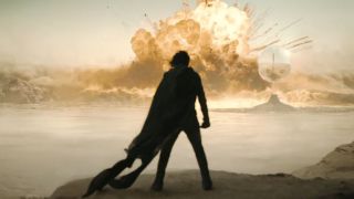 a man in cloak stands in the desert watching a massive explosion in the distance