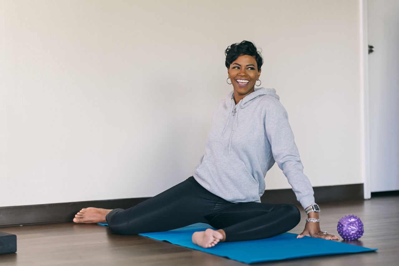 A woman on a yoga mat performs a hip mobility move. Both her legs are bent at right angles and resting on the floor; one is in front of her and the other is out to the side. Her hands rest on the floor and she is smiling.