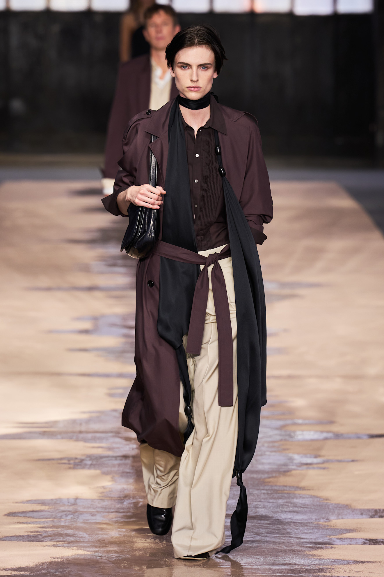 Layering staples for spring 2026 are shown in a photo from Boss's runway show, with a model walking while wearing a dark brown trench coat over a brown button-down shirt styled with a long black skinny scarf, tan wide-leg trousers, black flats, and a black shoulder bag