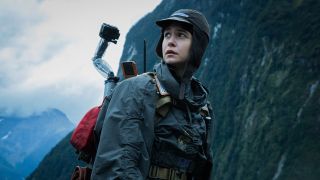 ill from the movie Alien: Covenant (2017). The image shows a person outside, standing in front of a mountain. They are wearing a jacket and a hat with flaps that go down past their ears. There is a red backpack on their bag with a robot poking out of it.