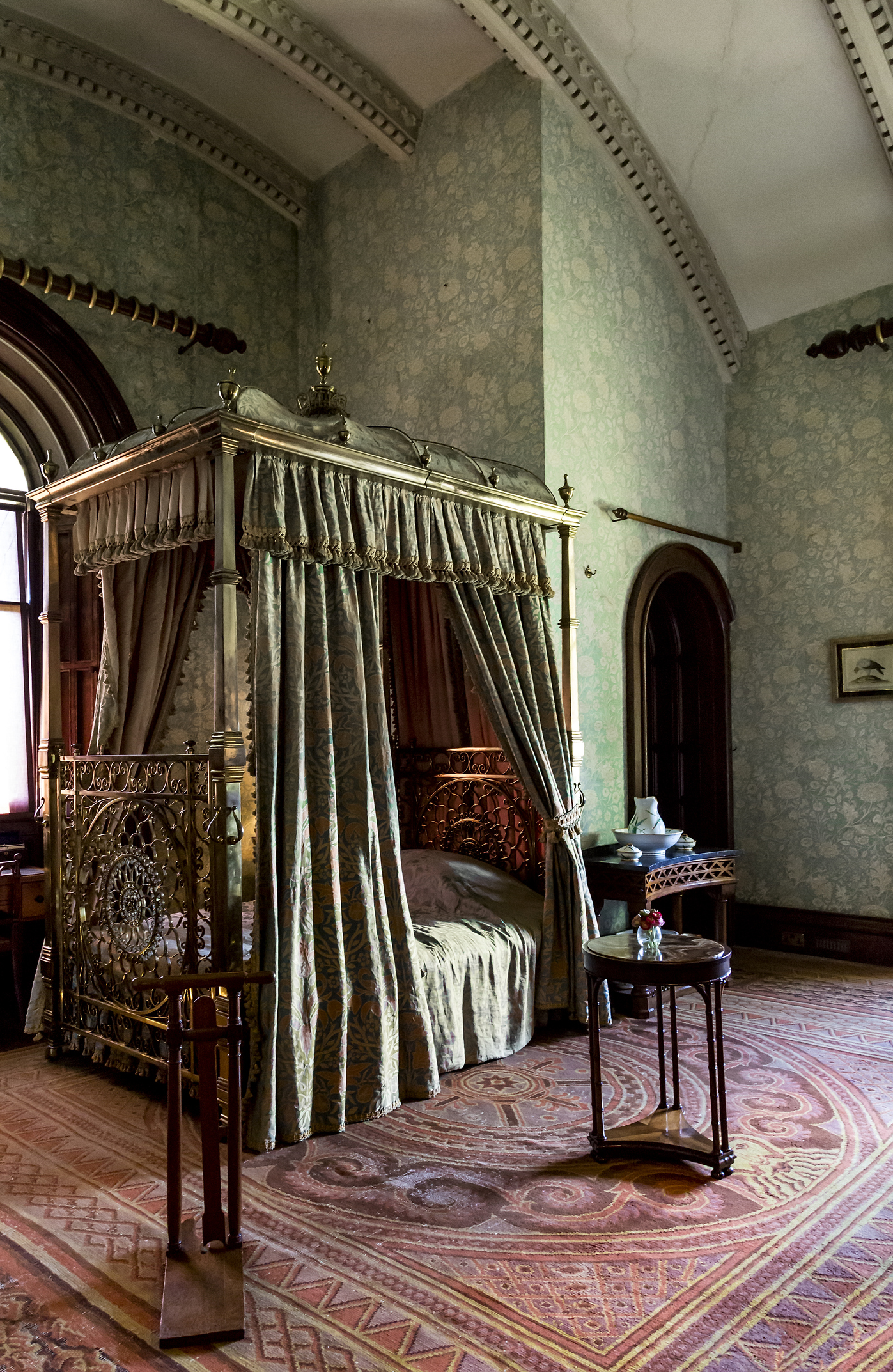 sage green bedroom in Victorian-era Penrhyn castle with heavy drapery, wallpaper, and red carpets, mahogany furniture, and tall ceilings