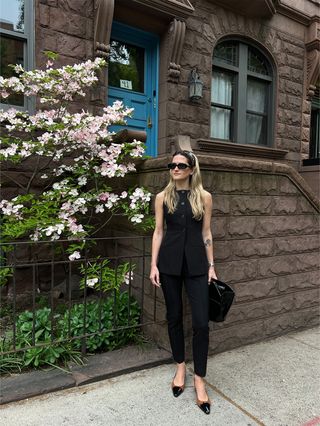 Eliza Huber mengenakan rompi hitam dan celana rokok dengan tas dengan pegangan atas berwarna hitam dan sepatu slingback berwarna coklat dan hitam.