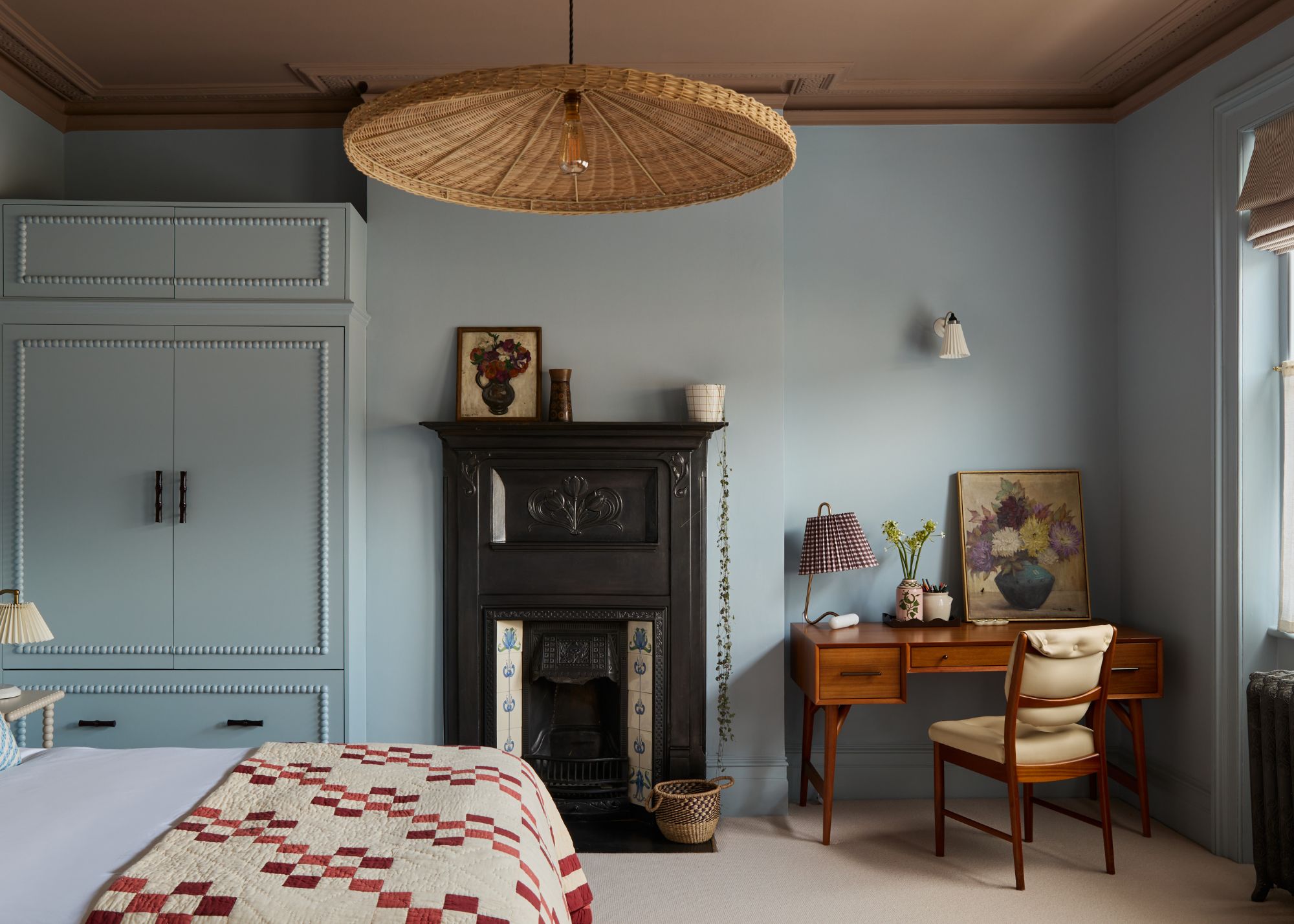 A calming bedroom with dusty blue color-drenched walls, a wooden desk and chair, a traditional black fireplace, taupe ceiling, a rattan ceiling light, and a bed with a red patterned quilt.