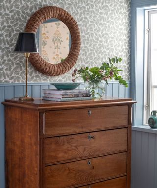 Close up of wooden dresser decorated with plants and books, round mirror above, blue painted paneling