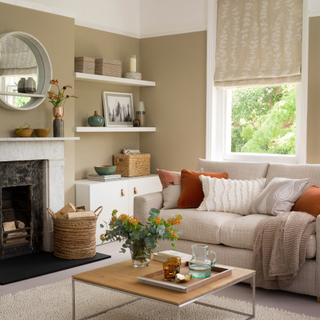 A cosy beige living room with orange and red cushions, borwn cofeee table, rustic fireplace and bouquet.