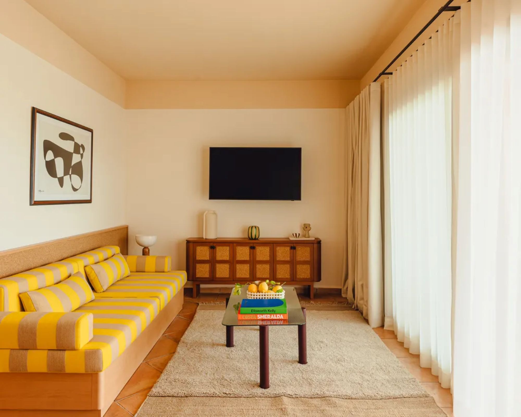 Yellow striped couch in neutral living room space with lemons on coffee table alongside colorful books. A TV rests on the back wall above a wooden credenza holding various decor objects