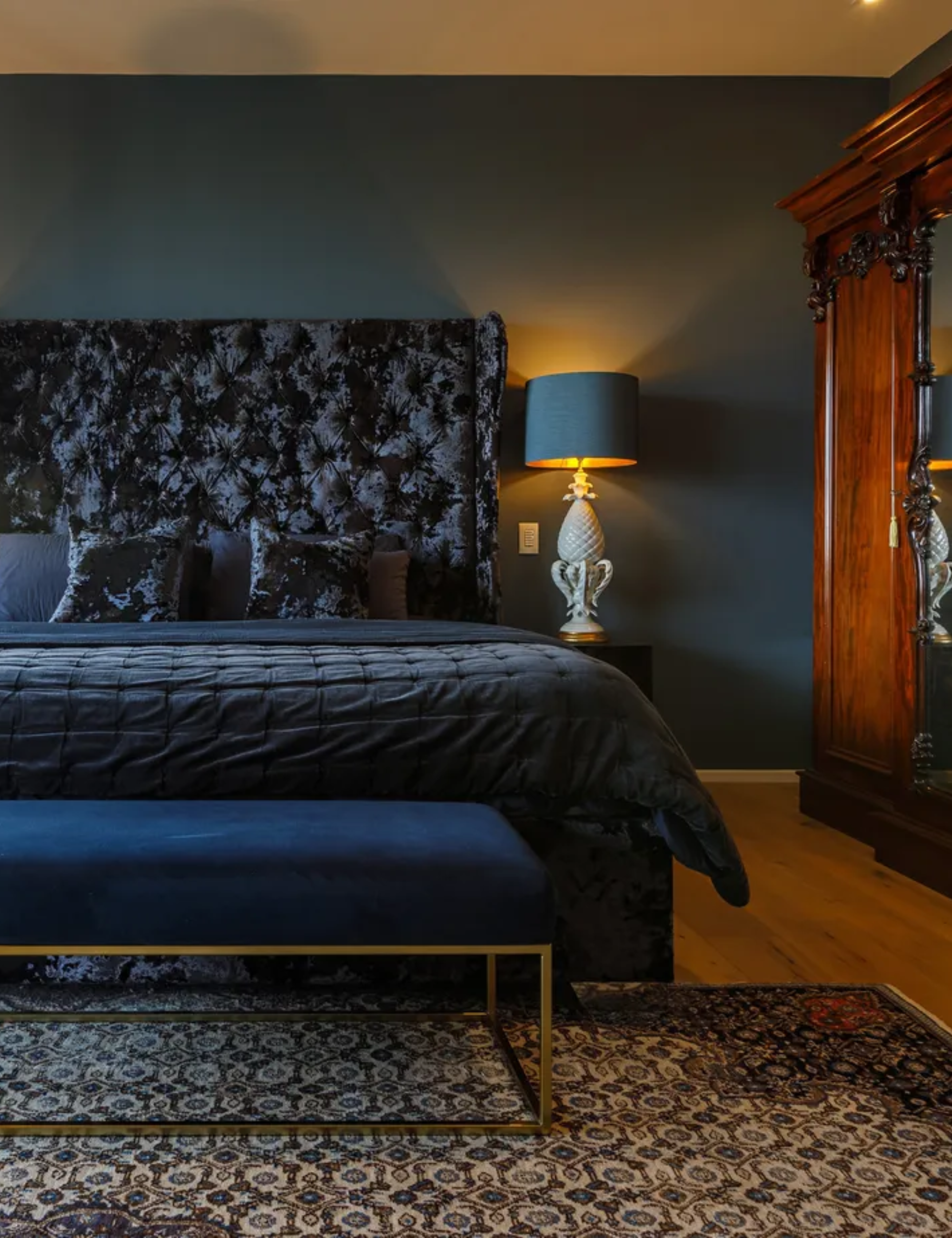 Dark bedroom, with large dark blue bed that has a tall, patterned headboard. A dimly lit lamp is on the bedside table, and an ornate wooden wardrobe is pictured to the right. A patterned rug and a footstool are pictured in front of the bed, on the floor.