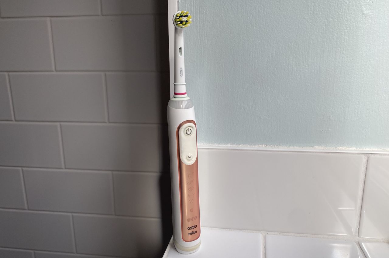 A rose gold Oral B Genius X electric toothbrush in a bathroom with white tiles and blue walls