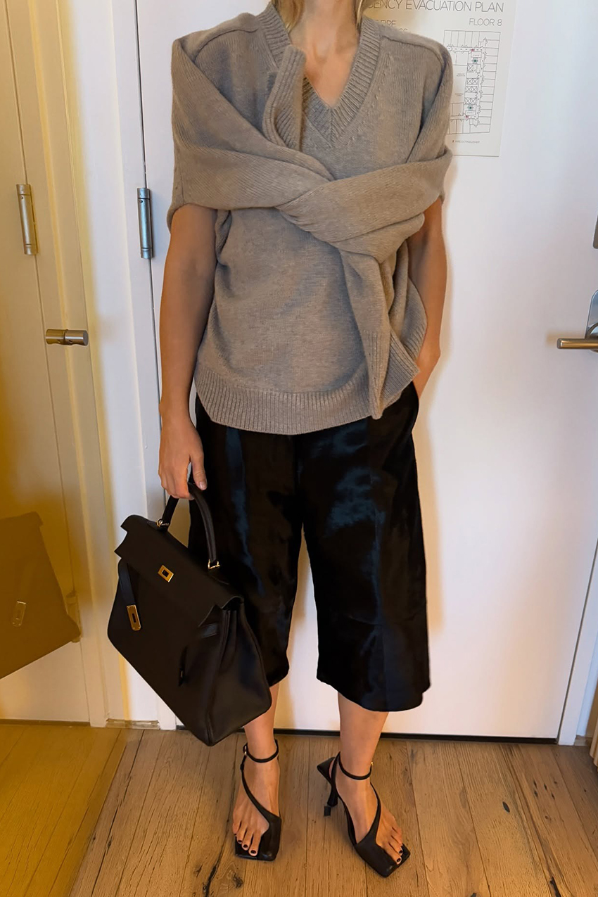 a woman wearing a beige sweater, black culottes, black strappy sandals, and a black leather bag