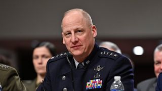 a man in a black military uniform sits at a desk behind microphones