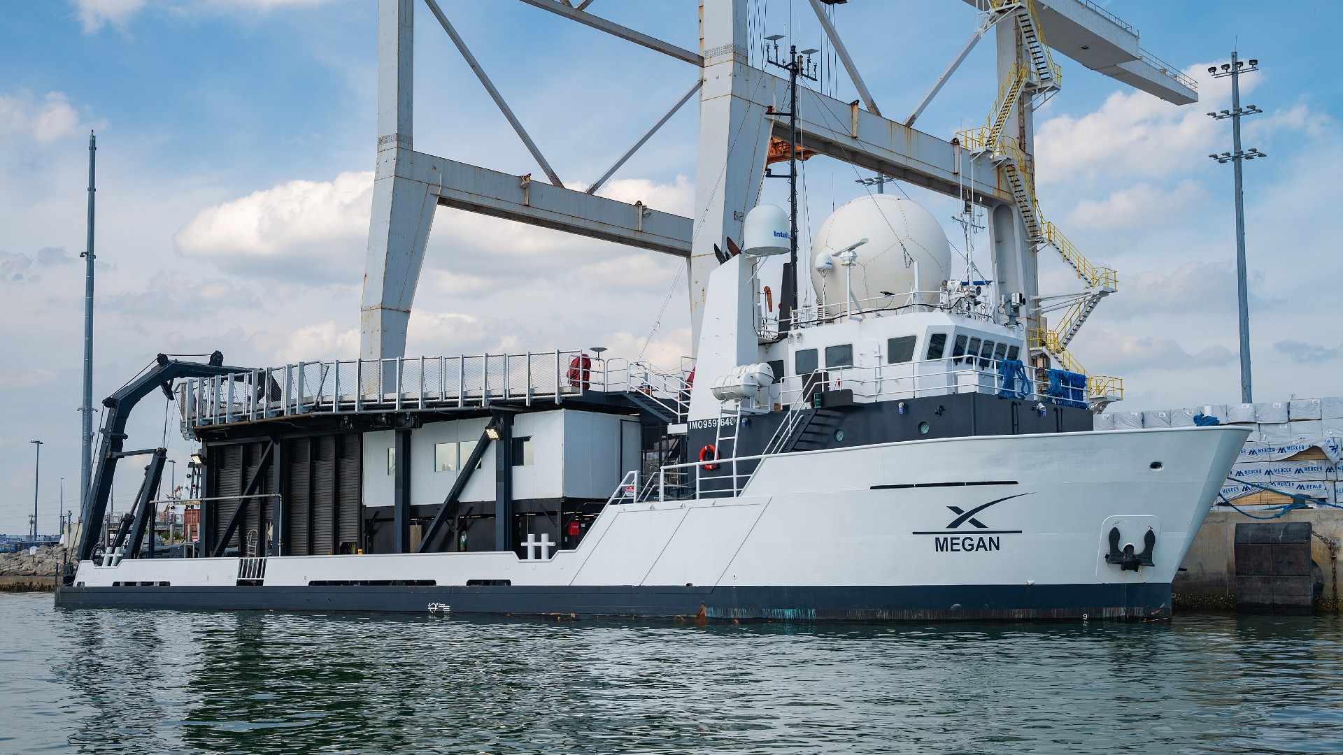 a large white vessel in a harbor. it features a wide, flat platform in the back and is emblazoned with the text "megan" under a large 'x'